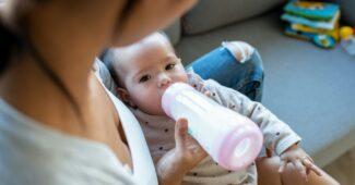 Mother feeding her baby daughter with feeding bottle while sitting on sofa at home.