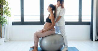 Physiotherapist helping pregnant for doing exercises with ball preparing for childbirth at home.